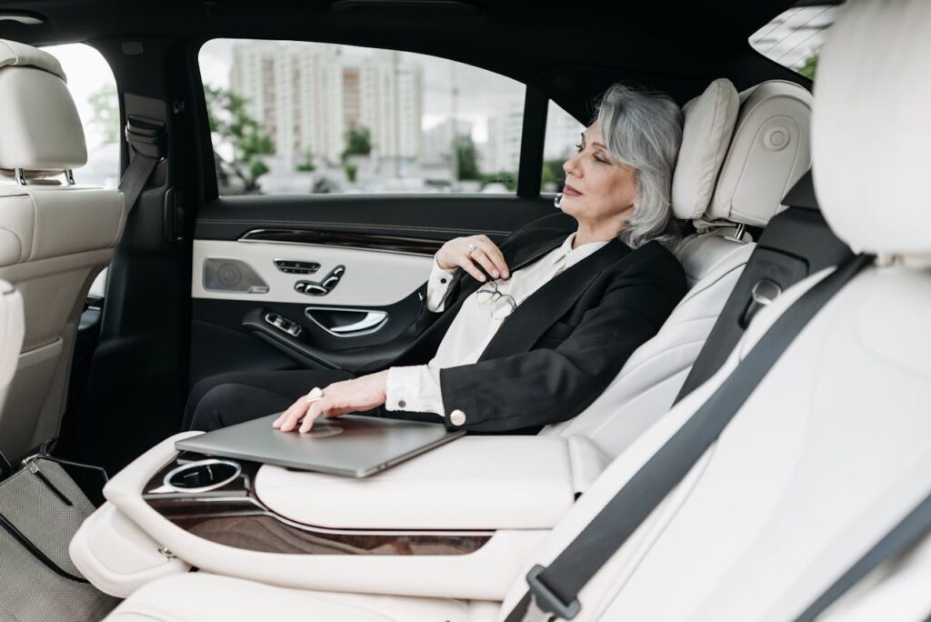 Elegant senior businesswoman relaxing in the back seat of a luxury black car with white leather interior, holding her glasses and laptop in a comfortable corporate travel setting.