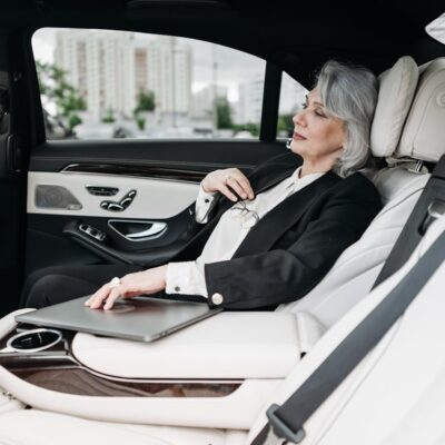 Elegant senior businesswoman relaxing in the back seat of a luxury black car with white leather interior, holding her glasses and laptop in a comfortable corporate travel setting.