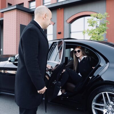 Professional chauffeur in a black suit and hat standing beside a luxury black car at a hotel, ready to assist arriving passengers.
