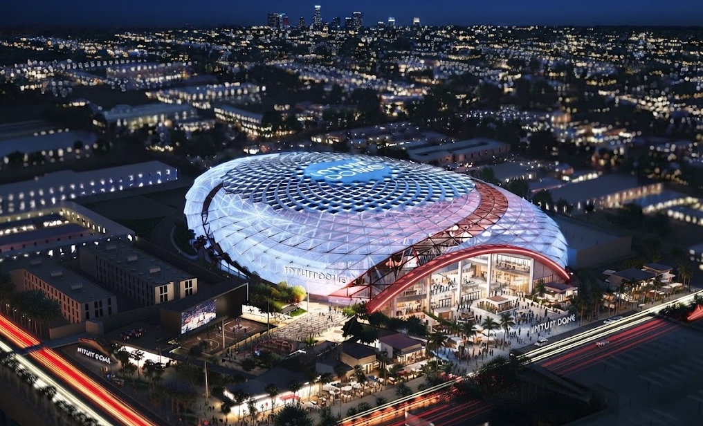 High-angle aerial night view of the illuminated Intuit Dome arena in Inglewood, surrounded by city lights and streaking traffic lines
